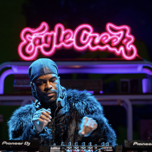 Image of madison moore standing at a DJ booth, with a pink neon sign behind them reading "Eagle Creek."